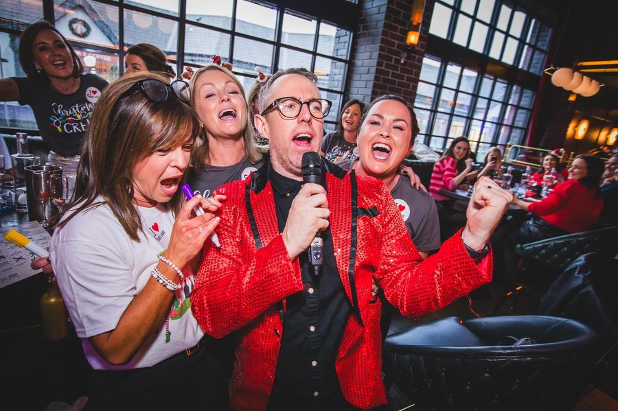 Patrick in a red sequin jacket on the mic at a Christmas party, surrounded by singing guests