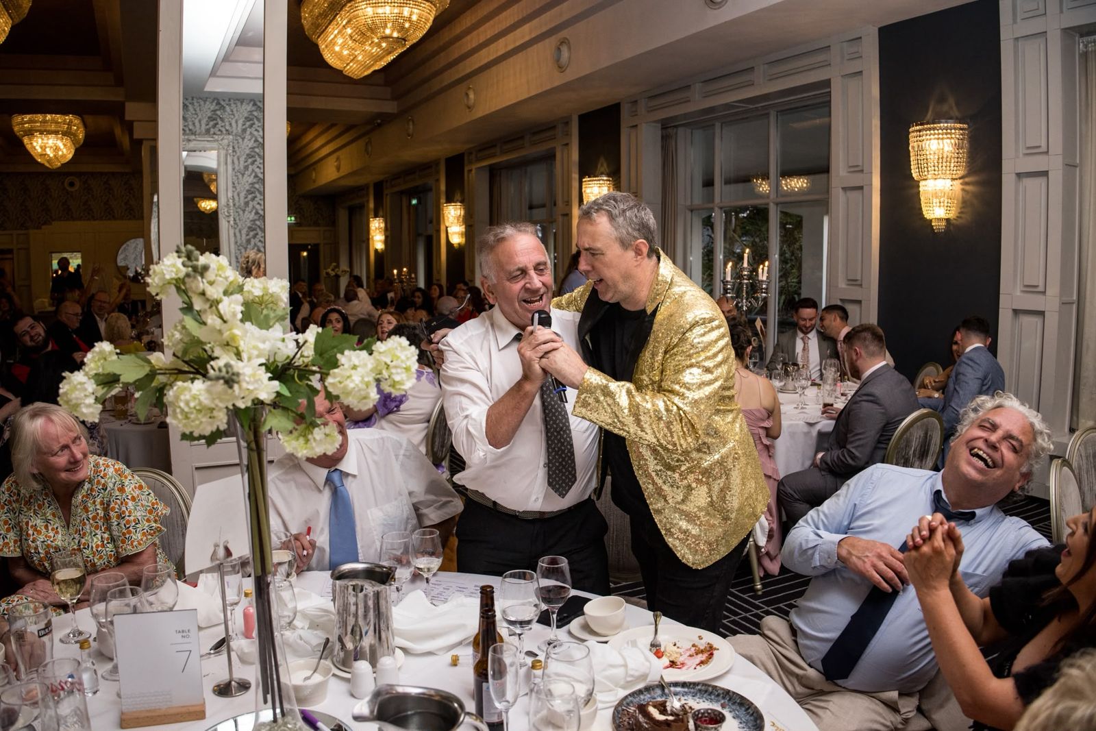 Patrick sharing the mic with a guest as the table laughs