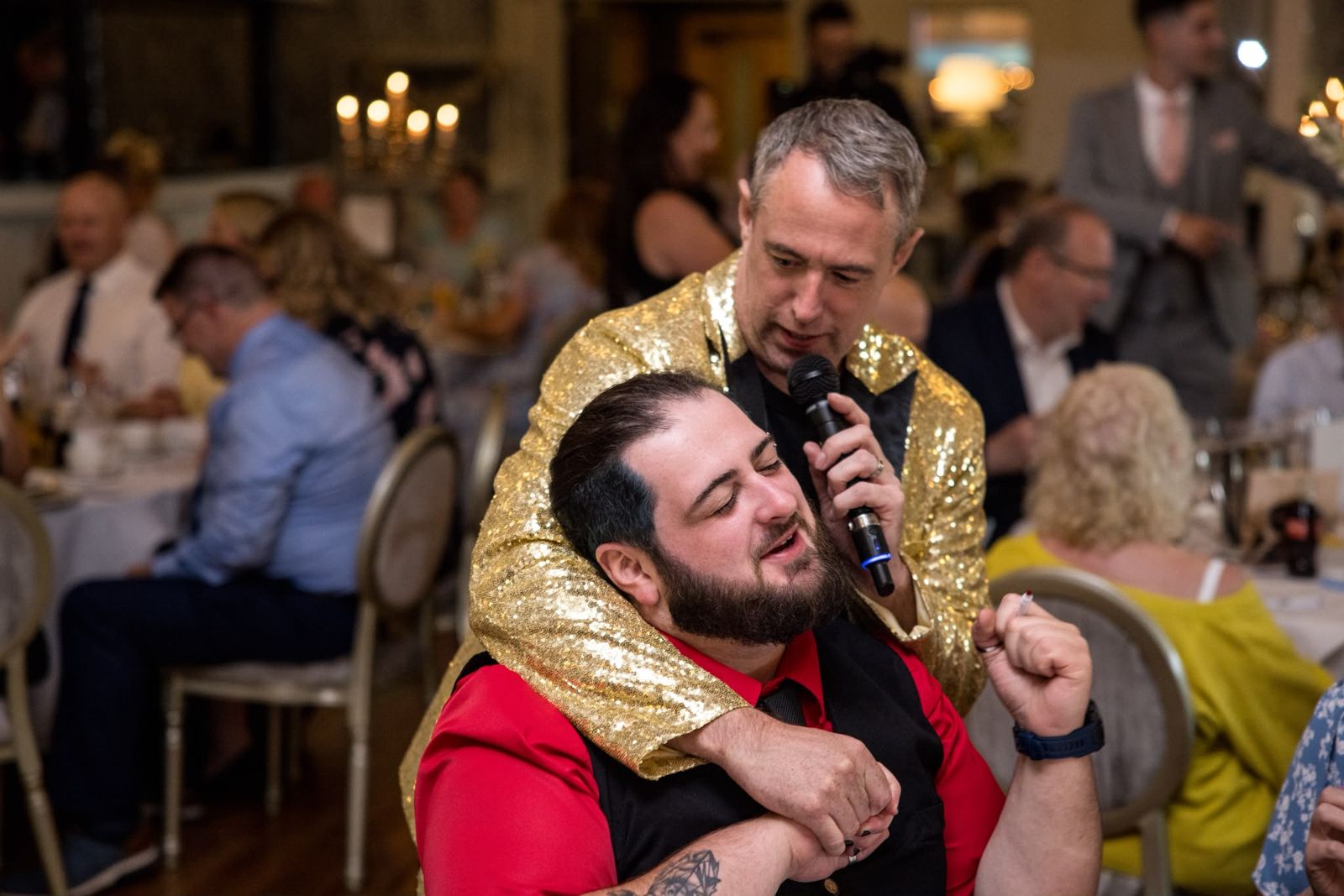 Patrick in a gold sequin jacket on the mic, arm around a guest singing at a wedding reception