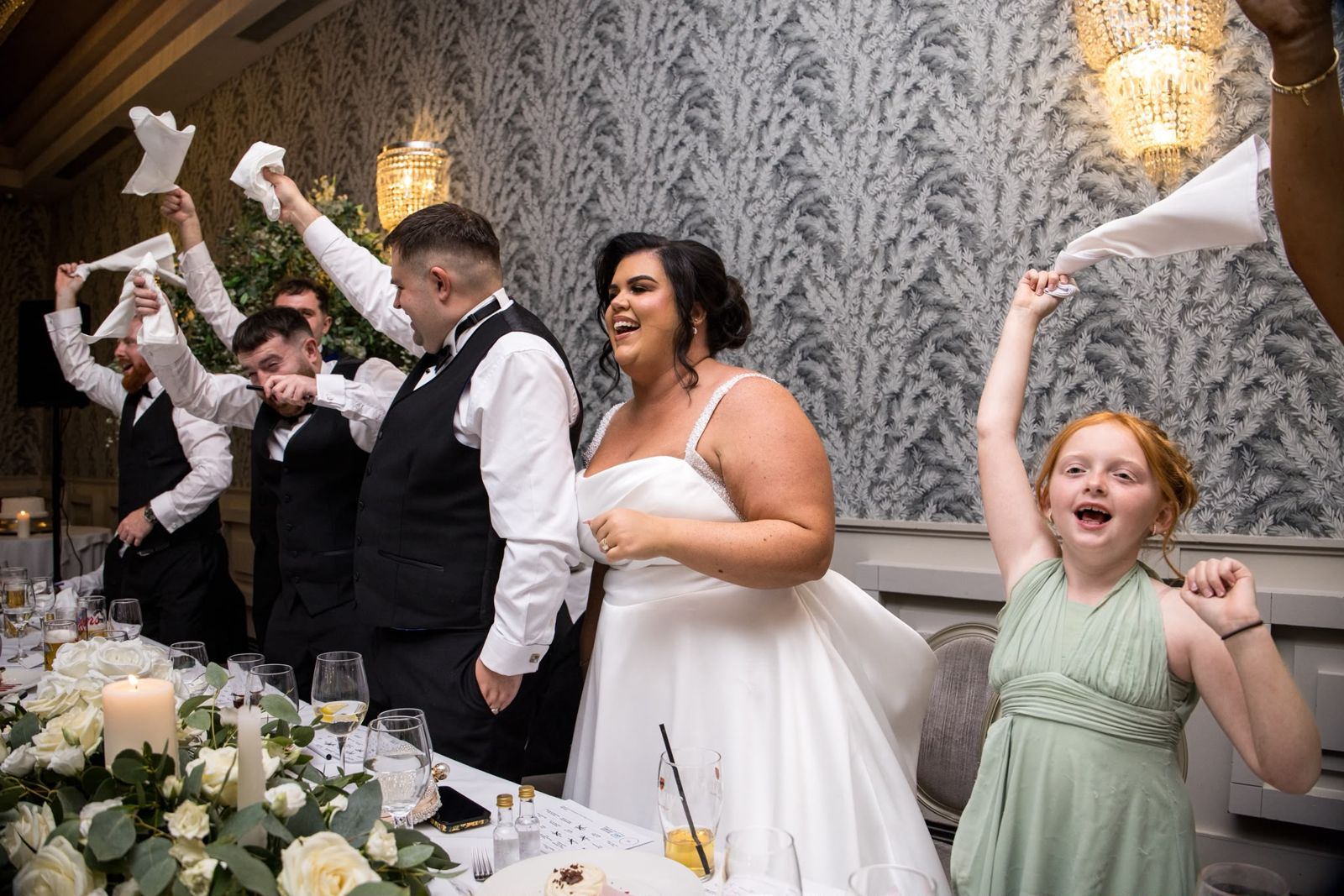 The bride, groomsmen and a young bridesmaid waving napkins over their heads at the top table